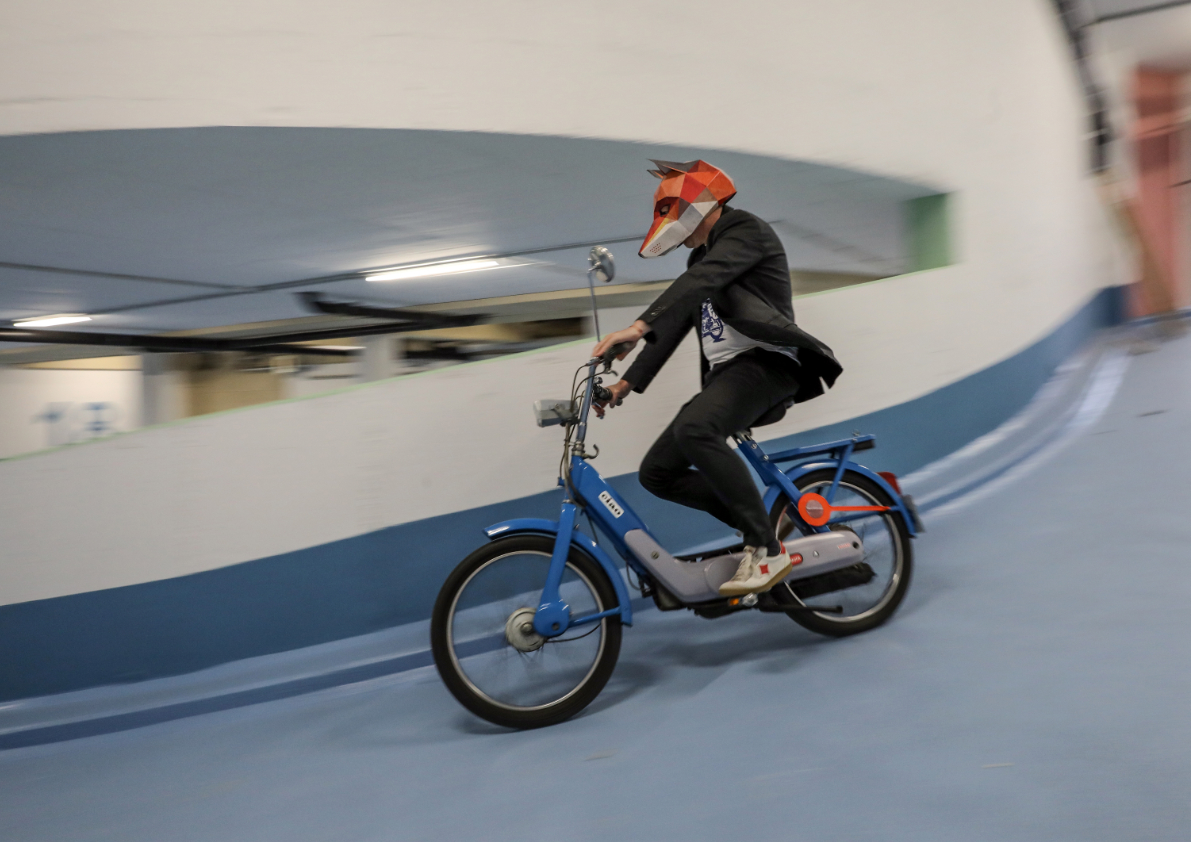 Person mit Fuchsmaske fährt auf einem Moped durch eine geschwungene Innenraumstruktur