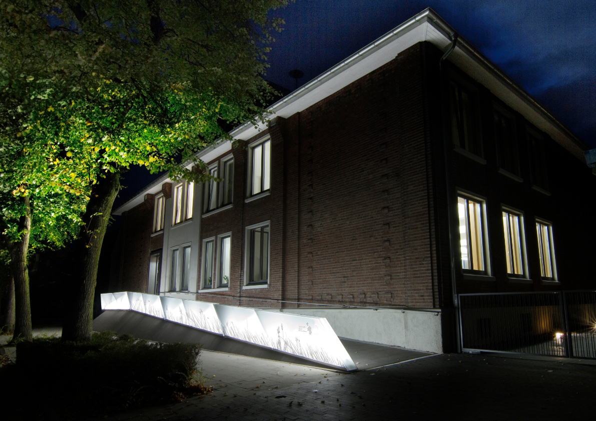 Schulgebäude bei Nacht mit beleuchtetem Sockel und Backsteinfassade unter Bäumen mit barrierefreiem Zugang.